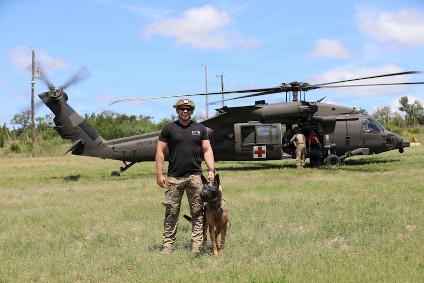 A man wearing a black T-shirt, camouflage military pants and helmet holds the collar of a dog as they pose for a photo while standing in front of a helicopter.