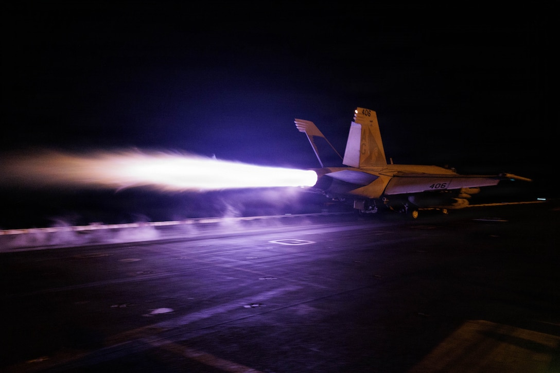 A fighter jet and its afterburner is seen as the jet takes off from the deck of an aircraft carrier at night.