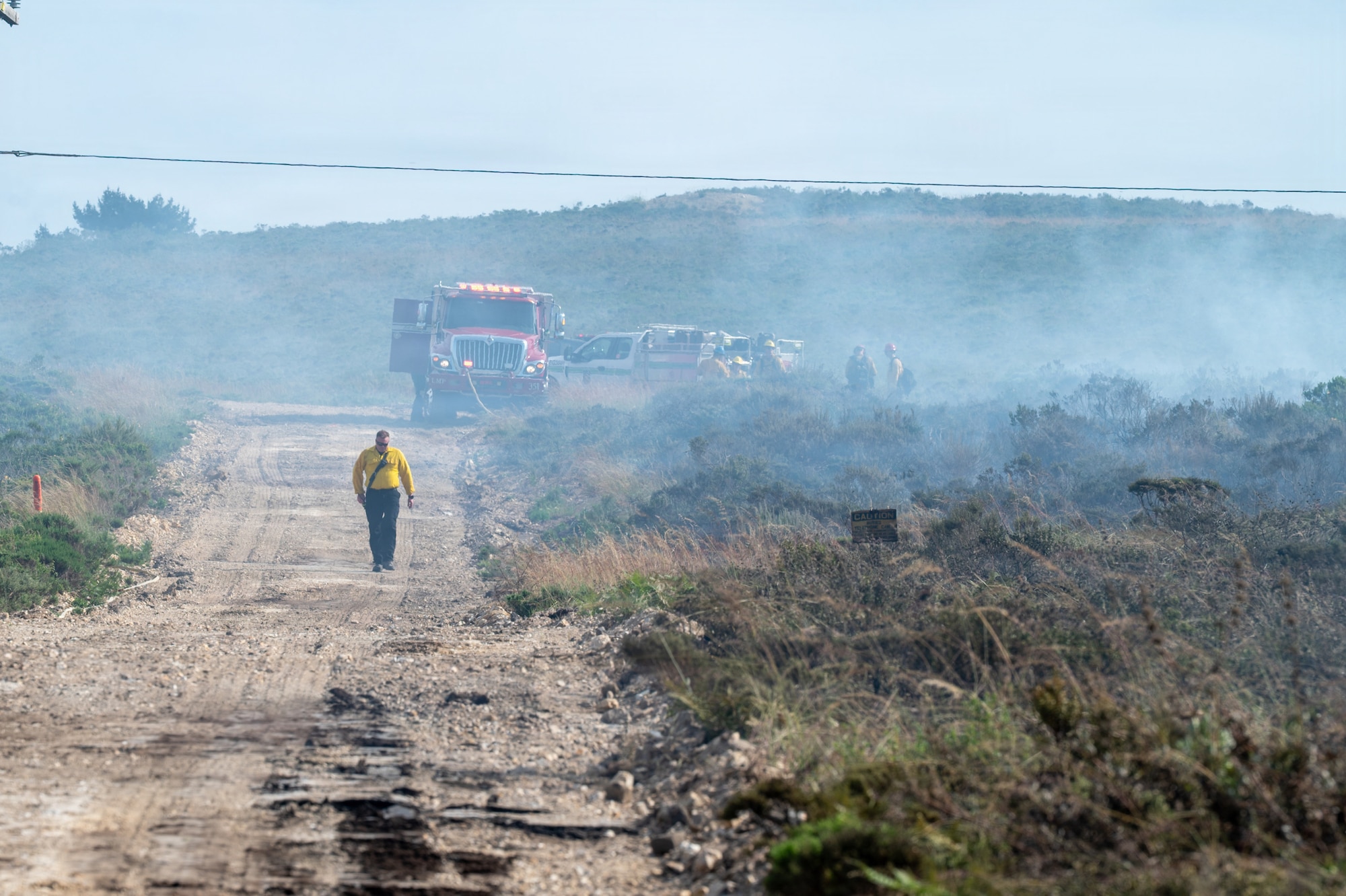 A man walks down a dirt road as smoke rolls in from the right. There is a firetruck at the top of the hill.
