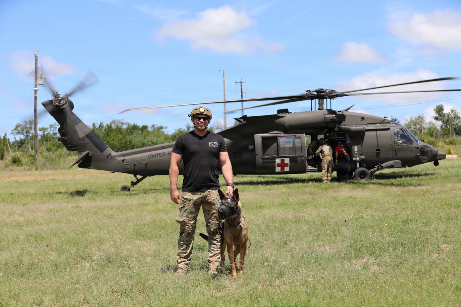 A man wearing a black T-shirt, camouflage military pants and helmet holds the collar of a dog as they pose for a photo while standing in front of a helicopter.