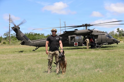 A man wearing a black T-shirt, camouflage military pants and helmet holds the collar of a dog as they pose for a photo while standing in front of a helicopter.