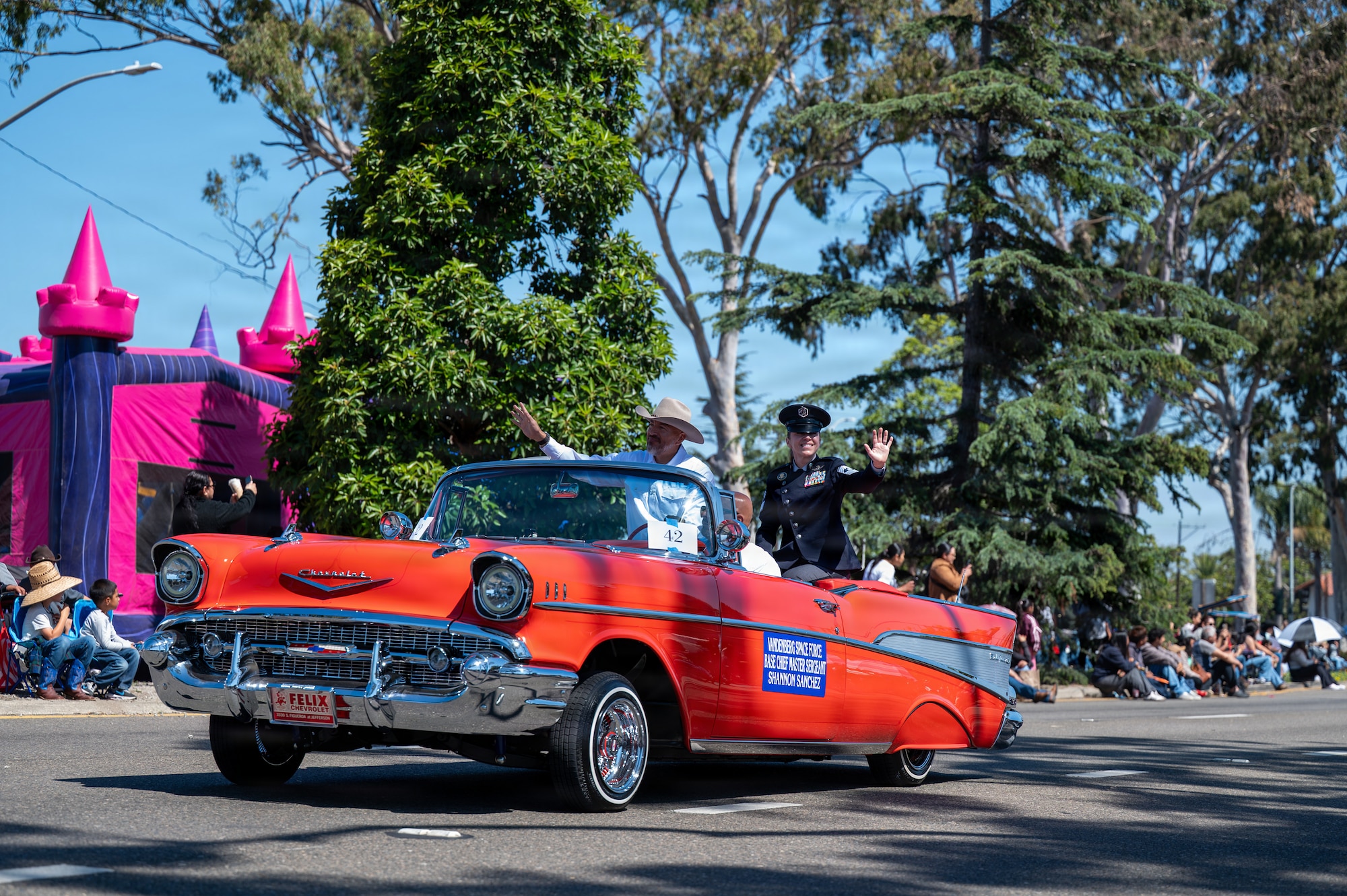 Orange car in the street with passengers waving during a parade.
