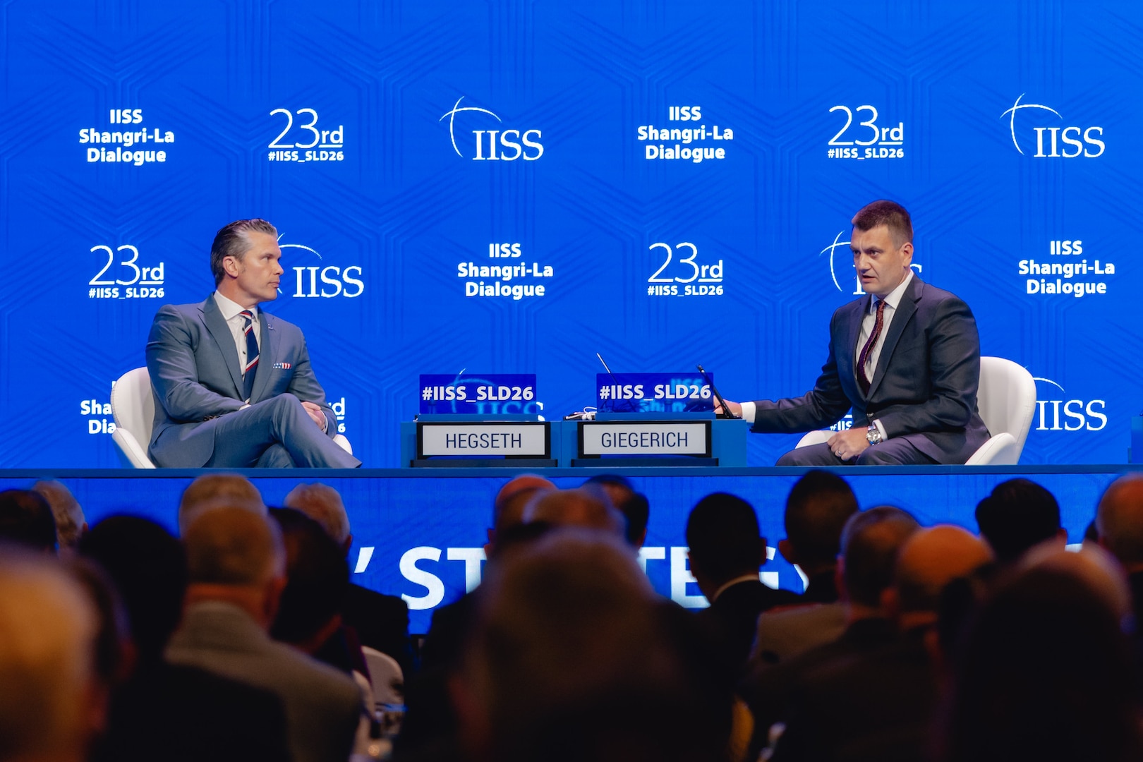Two men in suits sit up on a stage against a blue background in front of an audience.