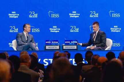 Two men in suits sit up on a stage against a blue background in front of an audience.