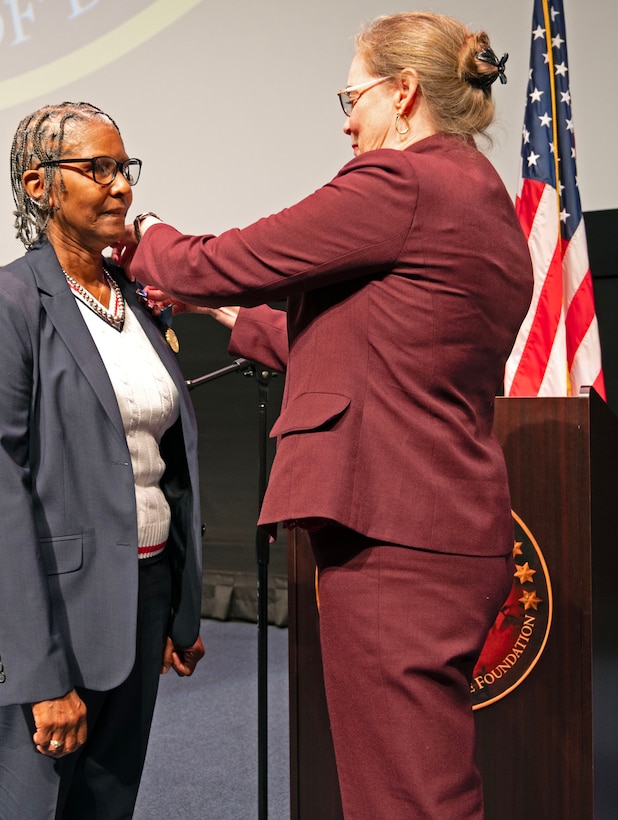 A smiling woman wearing a burgundy suit pins an award on a woman wearing a gray suit
