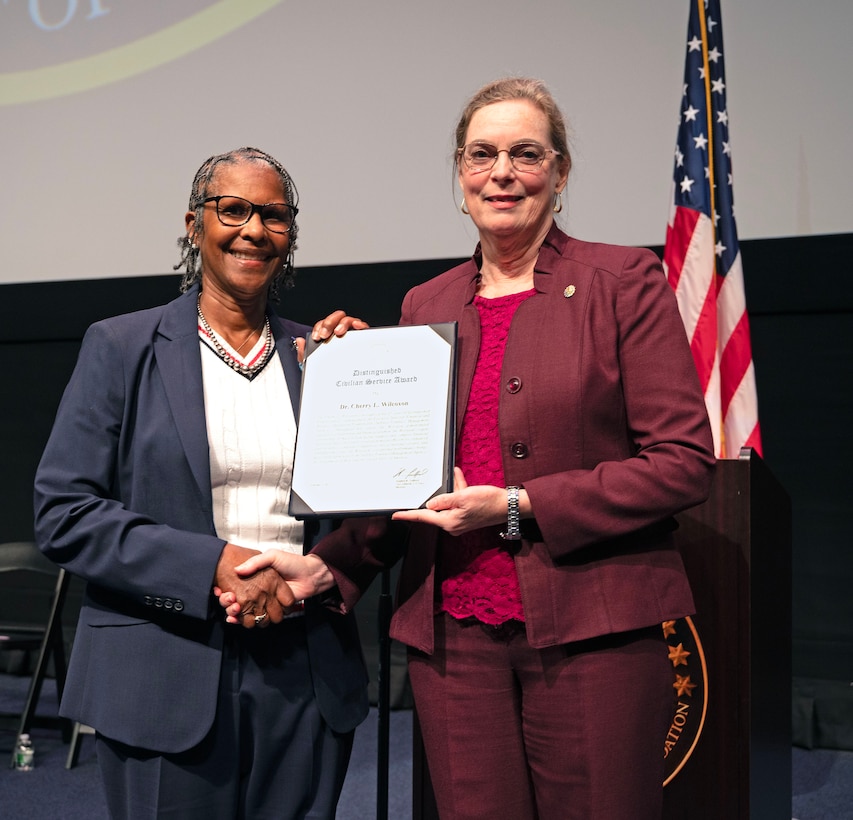 Two smiling women hold a certificate and stand near the American flag