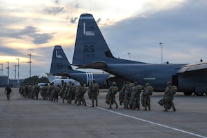 Dozens of people wearing camouflage military uniforms and parachute gear walk toward military aircraft parked on a tarmac.