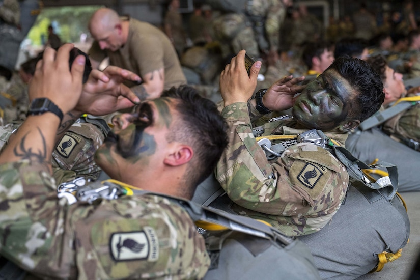 Two men wearing camouflage military uniforms put camouflage paint on their faces while lying in an aircraft hangar.