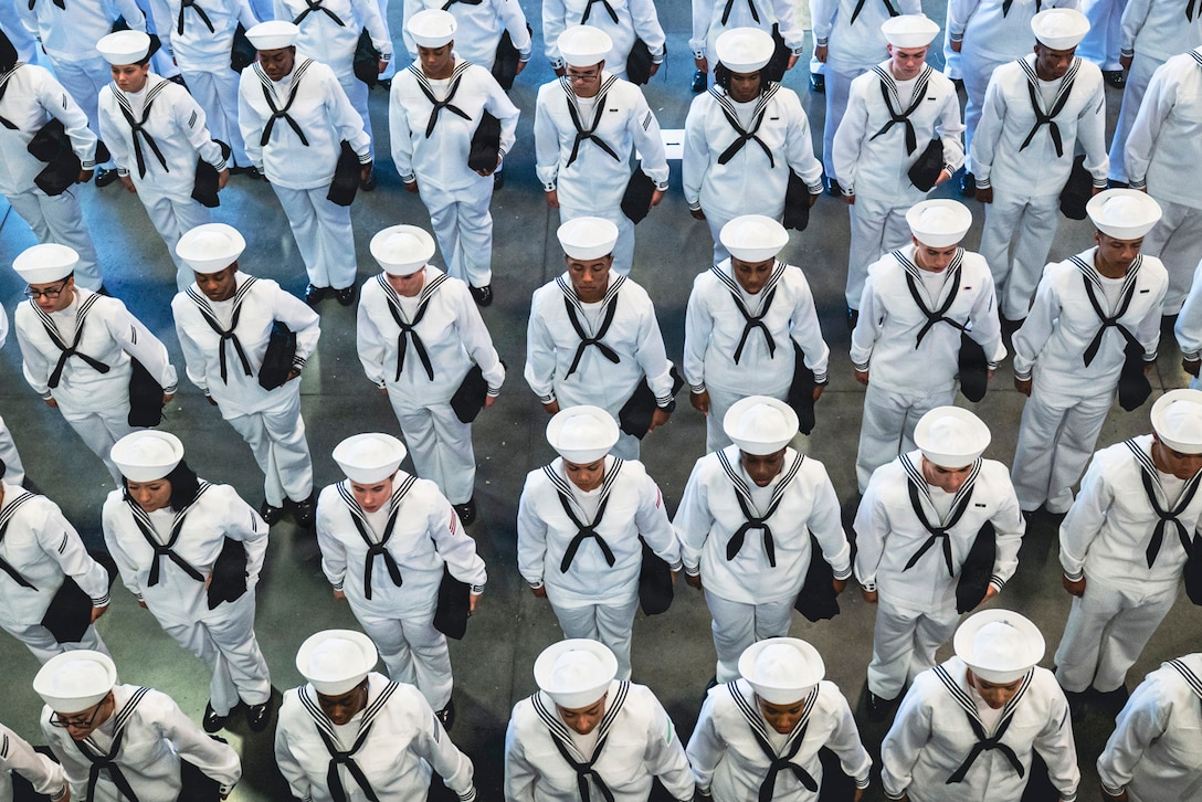Sailors in white and navy uniforms stand in formation while holding a black case as seen from above.