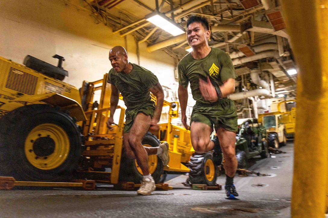 Two Marines strain while racing up a ramp near parked construction vehicles in a dimly lit maintenance room.