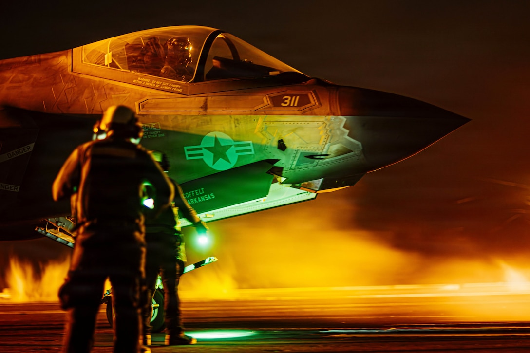 Sailors holding neon batons in the dark walk toward an aircraft on a ship surrounded by smoke.