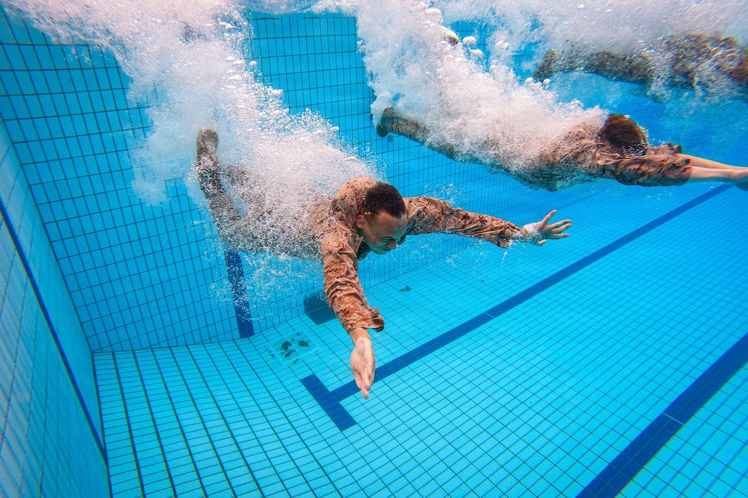 Marines in uniform make a splash while swimming underwater in a pool.