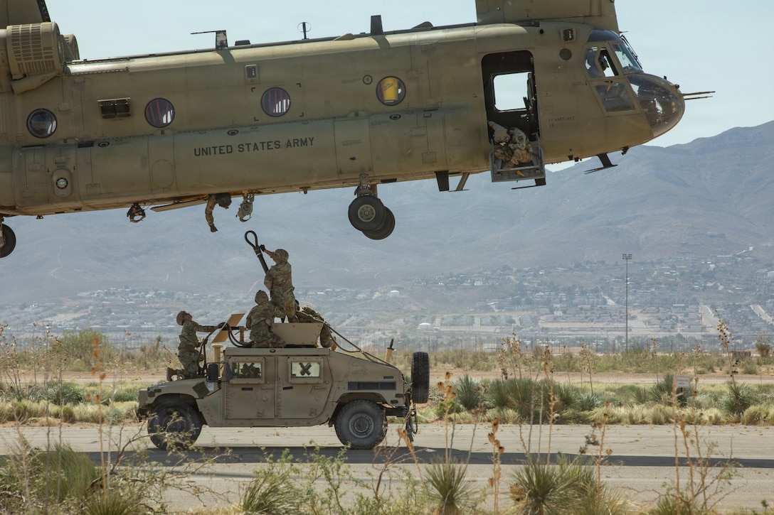 Three soldiers wearing camouflage uniforms and helmets kneel on top of a military vehicle and attach a cable connected to the vehicle to a helicopter hovering over them in a desert environment on a bright day.