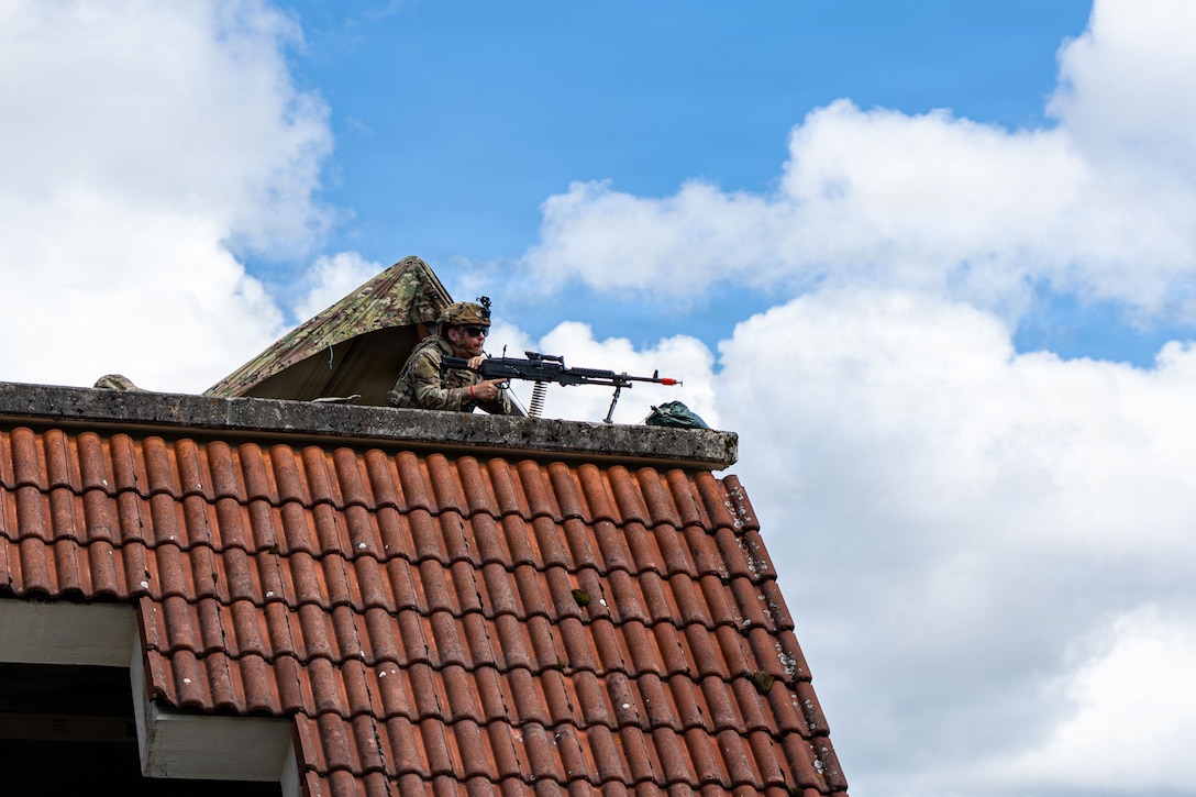 A soldier wearing a camouflage uniform and helmet lies in the prone position on the roof of a building while holding a machine gun on a bright day.