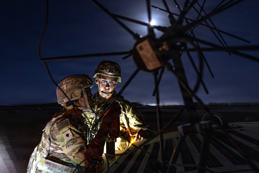 Two soldiers wearing camouflage uniforms, tactical vests and helmets set up a communications system at night outdoors under moonlight.