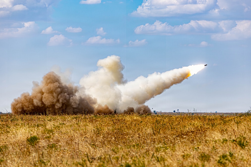A rocket produces fire and smoke as it takes off from a launcher on a semi-desert grass plain during a bright day.
