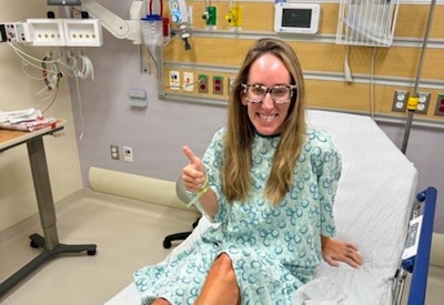 A woman wearing a hospital gown gives a thumbs up and smiles while sitting in a hospital bed.