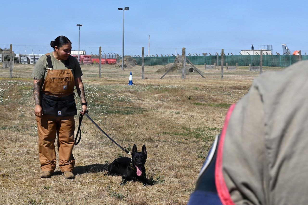A woman wearing brown overalls holds a black dog on a leash.