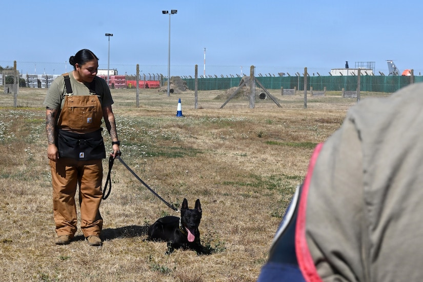 A woman wearing brown overalls holds a black dog on a leash.