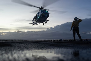 A helicopter takes off from the deck of a ship. A uniformed person salutes the helicopter.