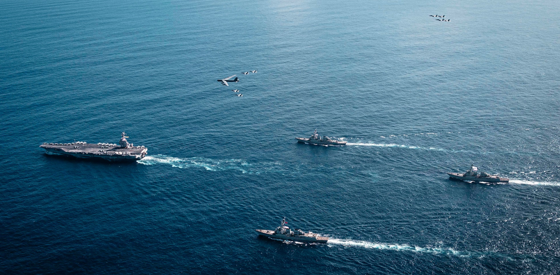 An overhead view of a group of four Navy warships steaming across the ocean in the daytime. A handful of military aircraft fly above them.
