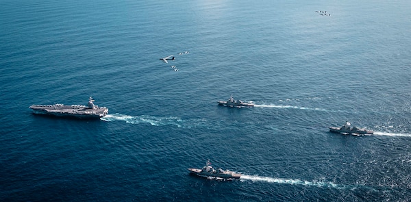 An overhead view of a group of four Navy warships steaming across the ocean in the daytime. A handful of military aircraft fly above them.