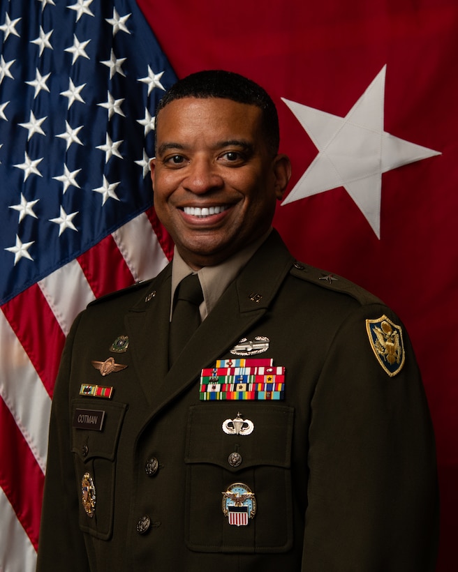 A man wearing a military uniform poses for a photo in front of an American flag and an Army one-star general flag.
