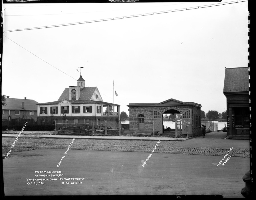 A view of two yacht club buildings on the Washington Channel waterfront. On the left is the two-story Capital Yacht Club, and to its right is a smaller, arched brick building for the Powell Yacht Club, with the Municipal Market partially visible on the far right.