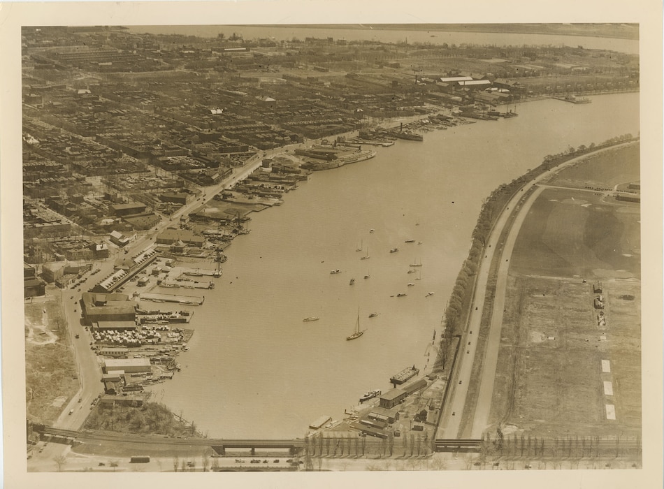 An aerial oblique photograph of a harbor and adjacent city. The left side shows a dense urban grid of streets and buildings; the right side features a broad roadway, tree-lined open land, and waterfront piers with numerous boats. The image documents overall conditions along the Potomac/Anacostia waterfront in 1926.