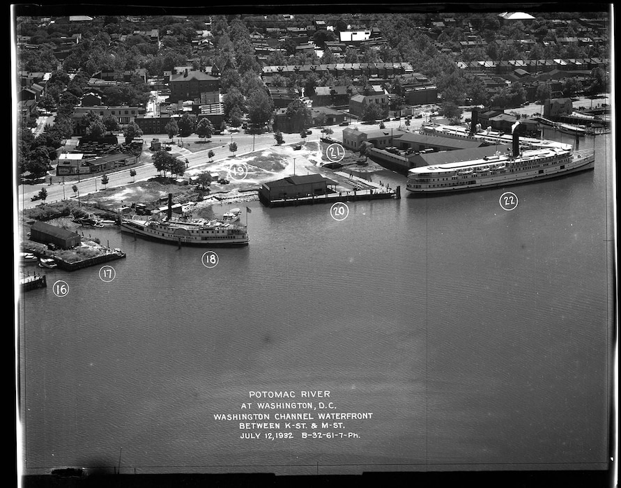 An aerial view showing two large passenger steamships docked at the waterfront. The ship at marker (18) is smaller than the large multi-deck steamer at marker (22). Numbered markers from (16) to (22). Businesses shown include: Capital Boat Works; Baltimore and Virginia Steamboat Co.; Steamer for Mt. Vernon. Vessels include: E. Madison Hall; ship of the N&W Line to Va. Seashore (Autos $3.00). Billboard posters advertise: NuGrape; Wonder Bread; tomato juice; Gulf gasoline.