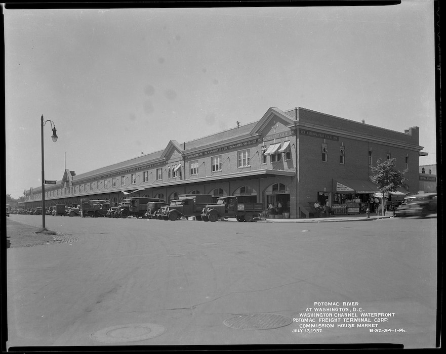 A street-level perspective of the long, brick Commission House Market building, part of the Potomac Freight Terminal. Numerous 1930s-era trucks are parked in front of the building's large arched loading bays. Angelo's Fruit Stand and Central Restaurant on the right. Some of the building's tenants are listed across the top.