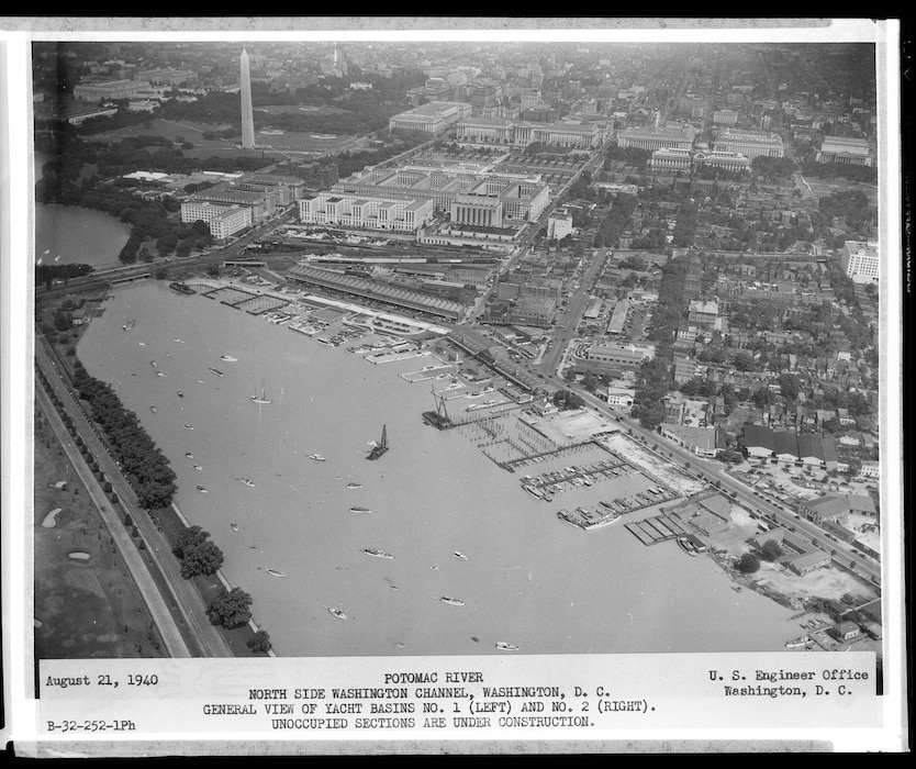 A high-altitude aerial view of the Washington Channel. The Washington Monument and the Federal Triangle are clearly visible. In the channel, a construction barge is visible, and many boat slips are empty, indicating ongoing construction.