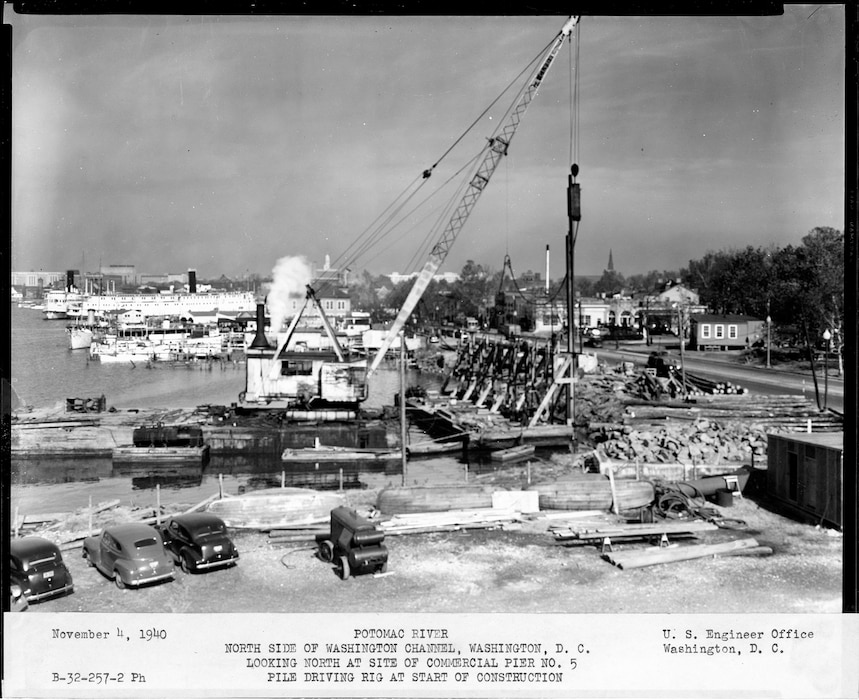 A wide view of the start of construction for Commercial Pier No. 5. Several cars from the 1930s and 40s are parked in the foreground. A large crane on a barge is operating in the channel.