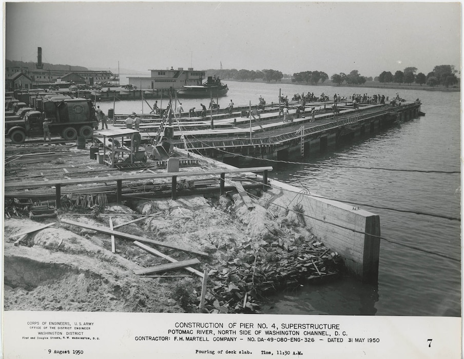 Workers and concrete trucks actively pouring concrete onto the deck slab, with wheelbarrows and reinforcing steel visible.