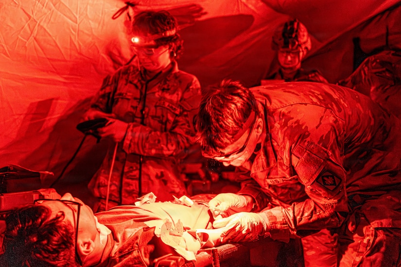 A man wearing a camouflage military uniform checks on a simulated patient in a tent at night; there are three other people in similar attire working in the background.