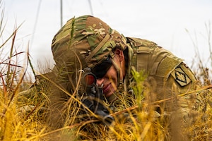 A man wearing a camouflage military uniform lies on the ground in a grassy field aiming a rifle.