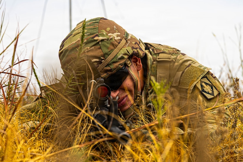 A man wearing a camouflage military uniform lies on the ground in a grassy field aiming a rifle.