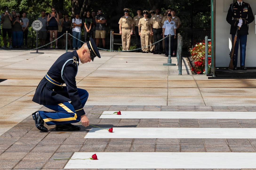 A soldier in ceremonial uniform kneels while placing a red rose on the concrete between other roses as fellow service members and civilians watch.