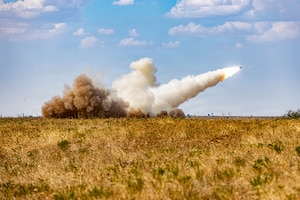 A rocket takes off from a field, shooting smoke and kicking up a cloud of dust behind it.