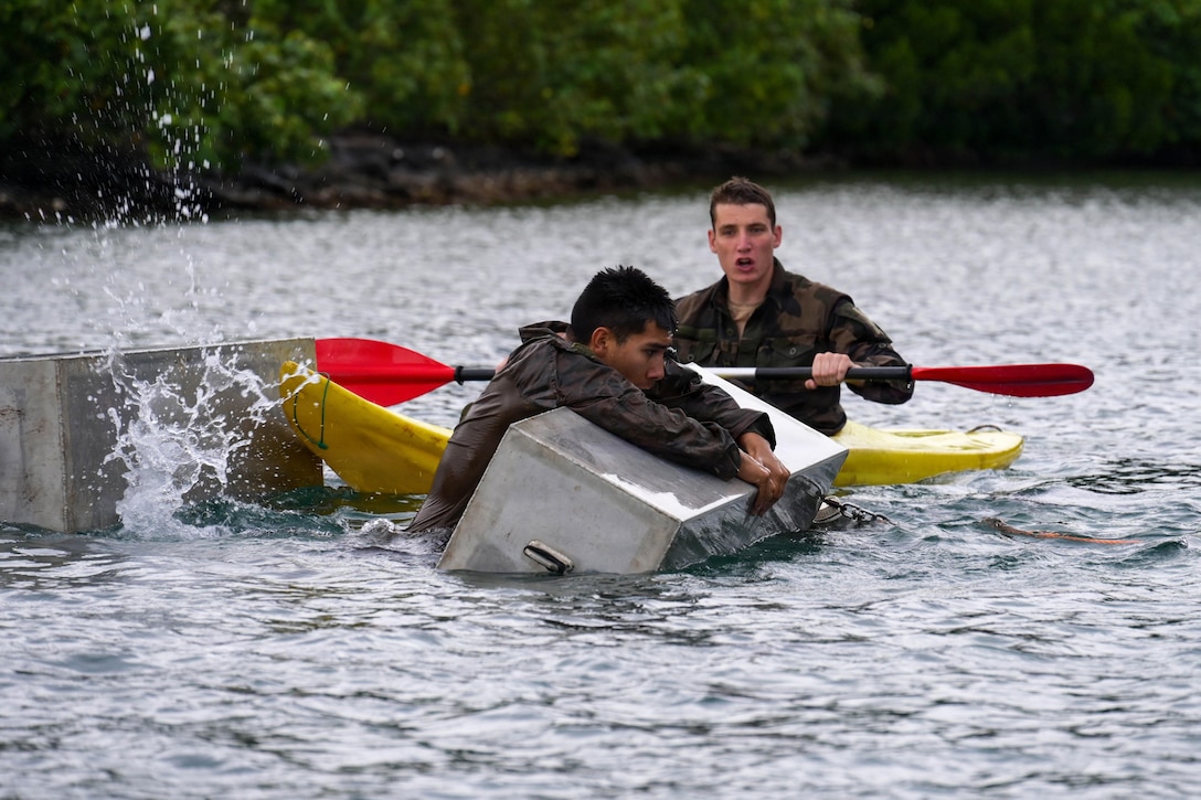 A Marine holds onto a barrier in a body of water as a fellow Marine in a kayak watches while holding an oar on a gloomy day.