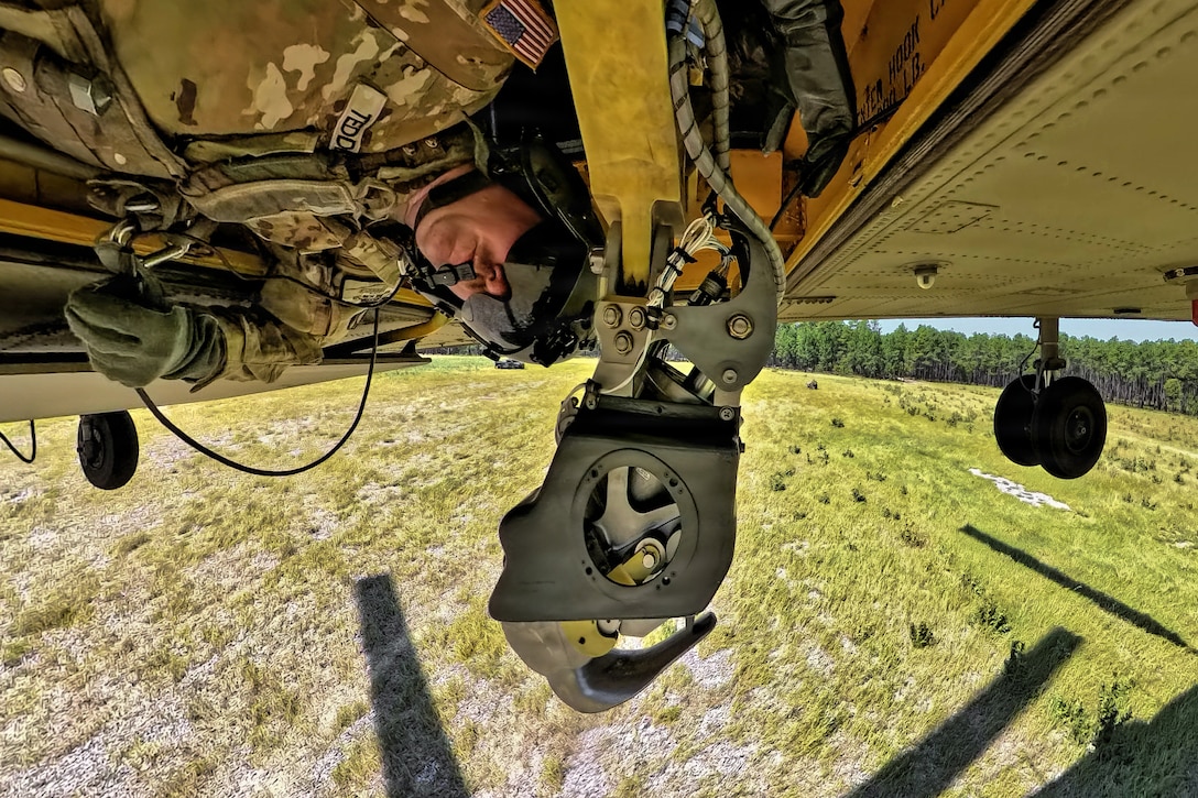 A soldier wearing a camouflage uniform and flight gear looks out from a portal on the underside of a flying helicopter lowering a hook on a bright day.