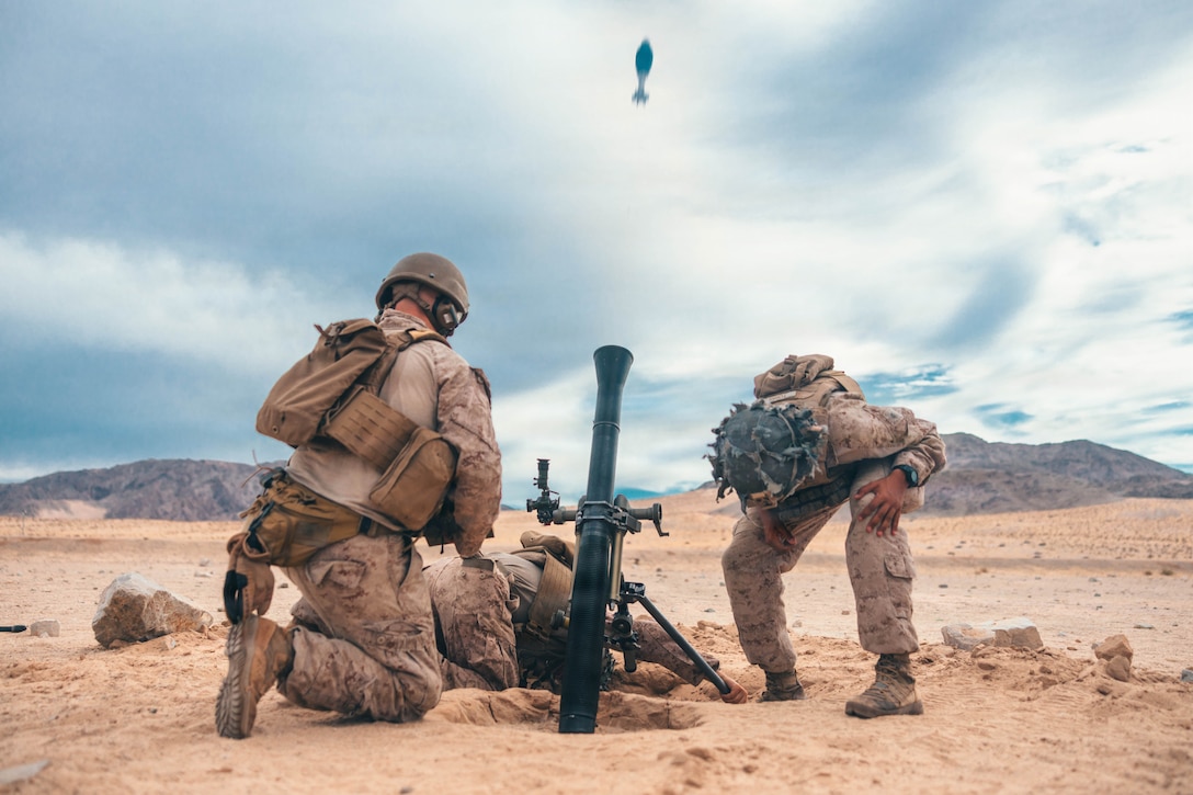 Three Marines wearing camouflage uniforms, helmets, and load-bearing equipment fire a mortar round out of a launcher in a desert environment on a bright day.