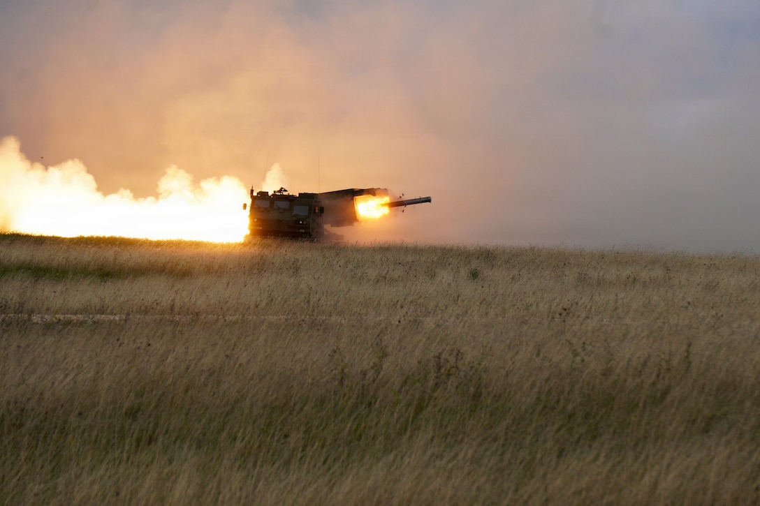 Flames erupt from a mobile rocket launcher as a rocket shoots out in a grassy field during daytime.