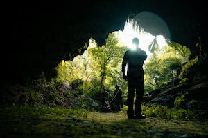 Daylight illuminates through a forest as it creates a silhouette of three men walking into a dark cave.