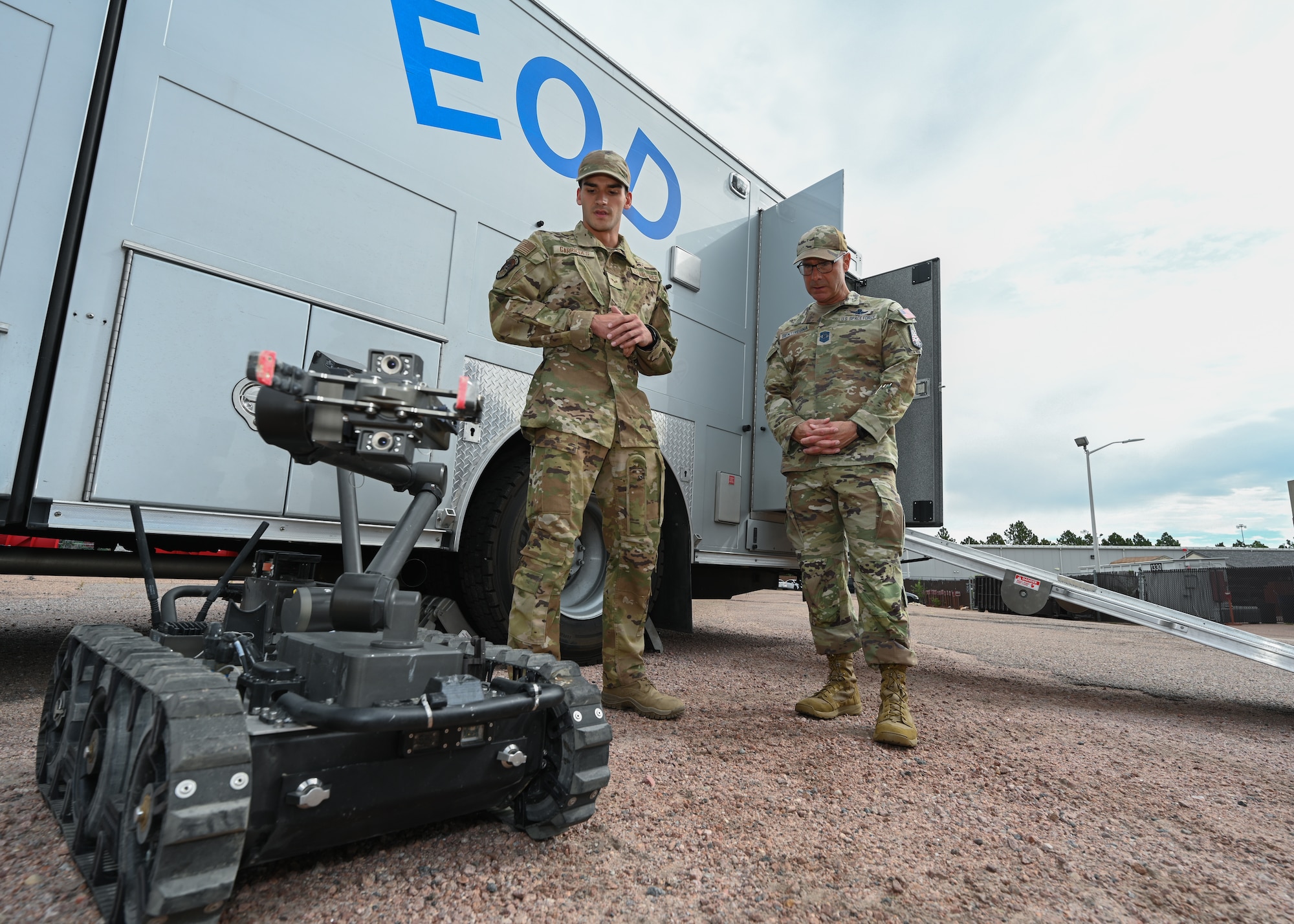 Both Chief Master Sgt. Bentivegna and Senior Airman Aaron Garrigues stand in front of a white EOD command trailer with a EOD robot next them.