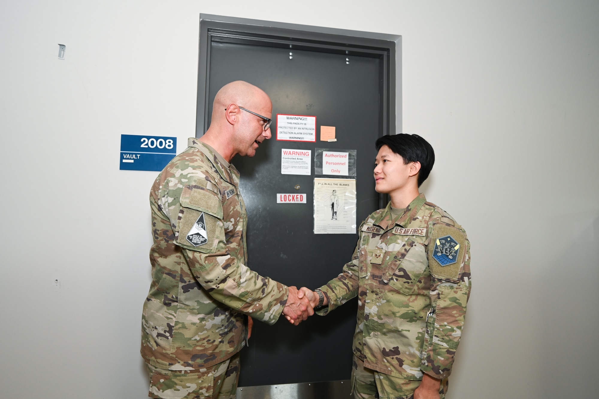 Chief Master Sgt. Bentivegna and Senior Airman Chloe Massenat stand in front of a door while shaking hands.