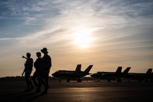 Three people in silhouette walk outside on a flight line, with three military aircraft parked behind them.