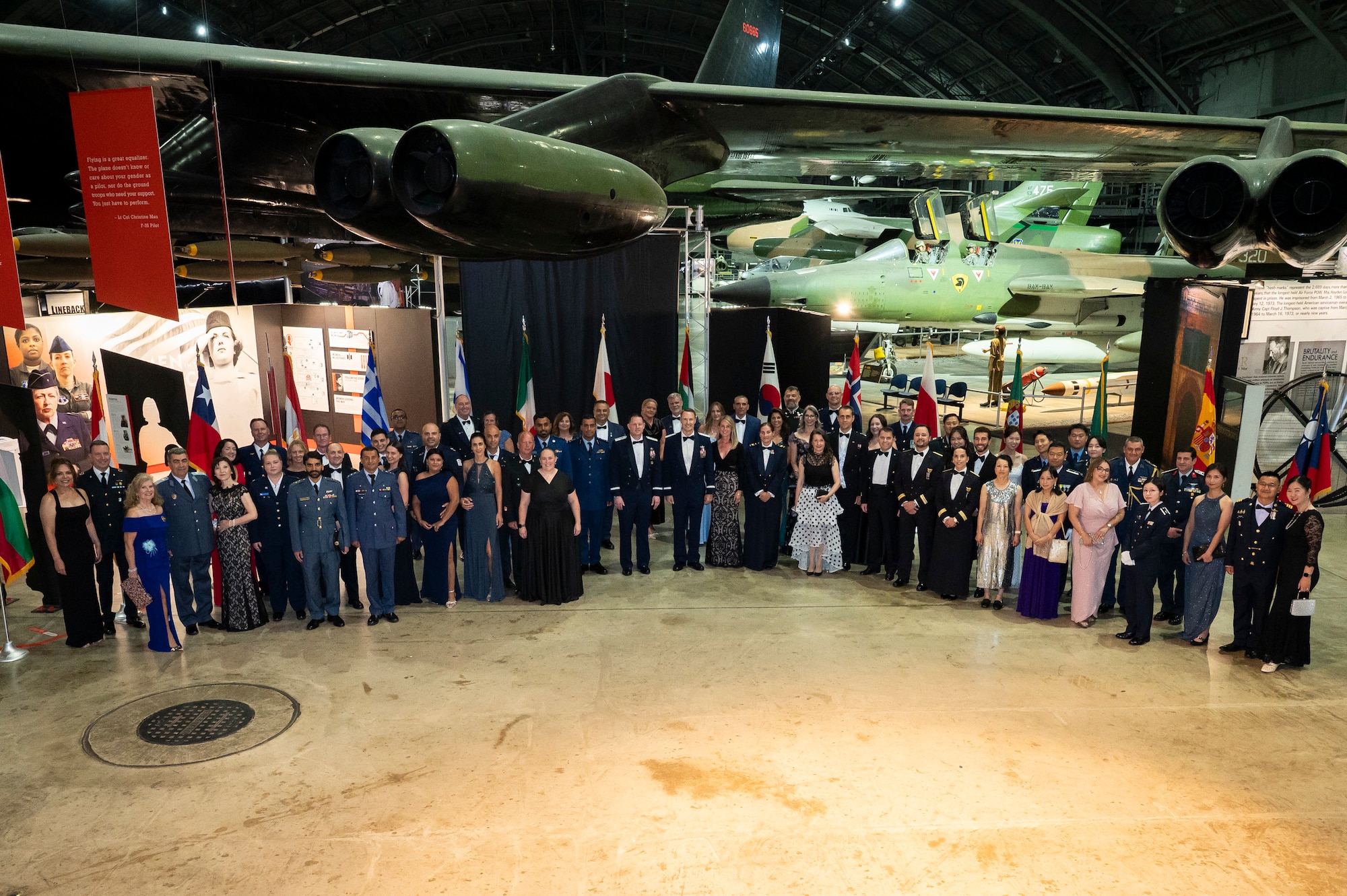 Large group shot of men and women wearing formal attire standing in a horseshoe shape posing for photo. Historical airplanes are in the background.