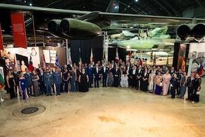 Large group shot of men and women wearing formal attire standing in a horseshoe shape posing for photo. Historical airplanes are in the background.