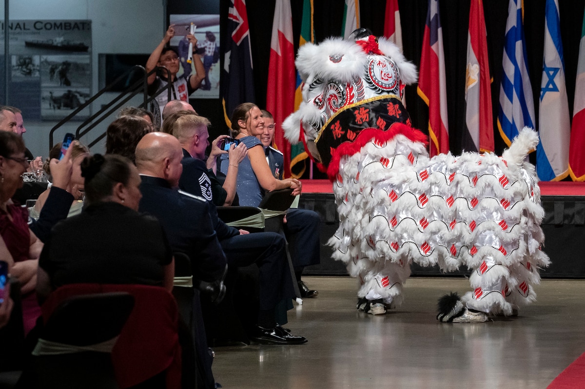 Person wearing dragon costume dancing. People at round tables. Red carpet is seen.